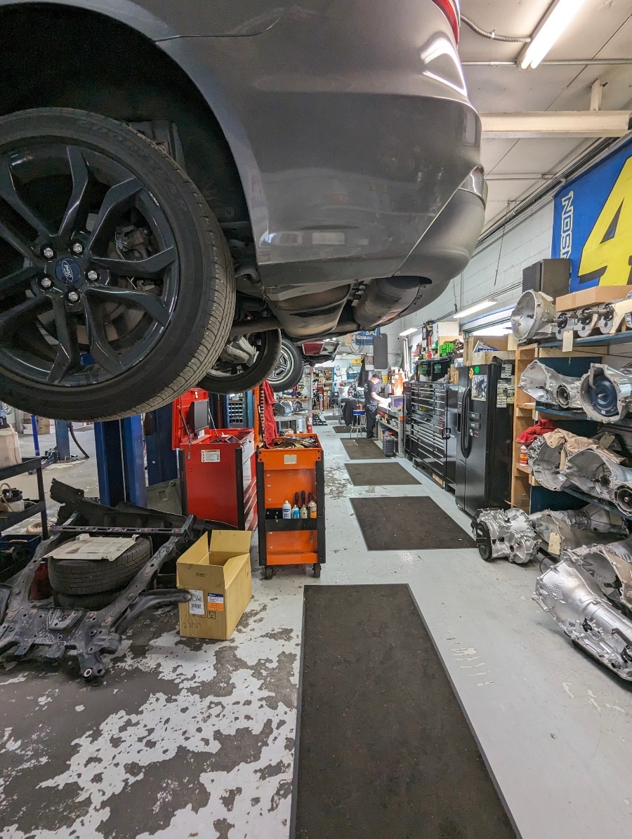 Under a lifted vehicle beside shelves of rebuilt transmission cases
